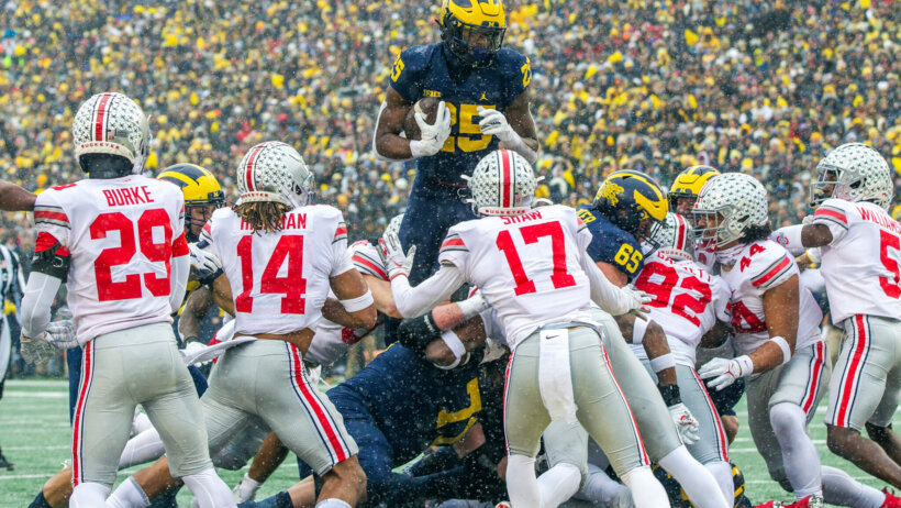 Michigan running back Hassan Haskins leaping over Ohio State players during a football game.